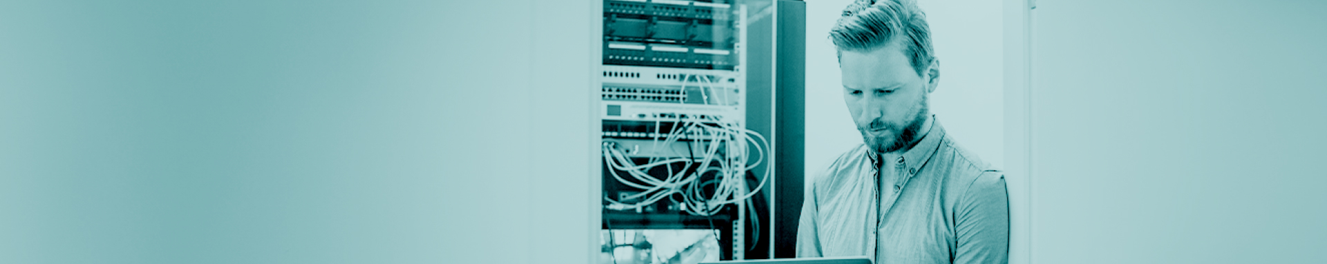Ein Mann steht angelehnt in einem Türrahmen vor einem Serverraum und arbeitet konzentriert an seinem Laptop.